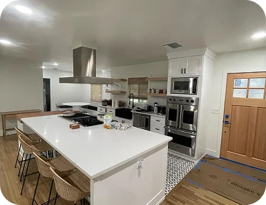 Spacious open-concept kitchen remodel by TG Renovations featuring a large white island with wicker seating, built-in stainless steel appliances, open wood shelving, and light hardwood floors.