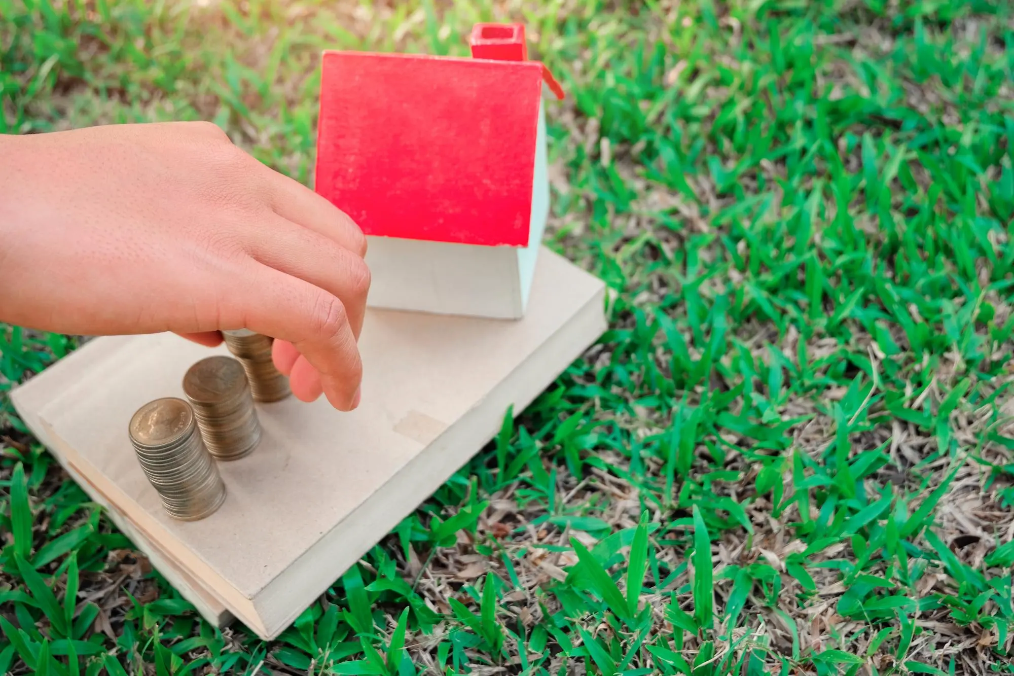 Hand stacking coins next to a miniature house model on grass, representing backyard home construction costs in Los Angeles – TG Renovations