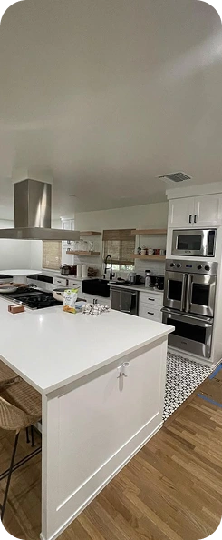 A bright, modern kitchen remodel by TG Renovations featuring a large white island, stainless steel double ovens, open wooden shelving, white cabinetry, and a black farmhouse sink.