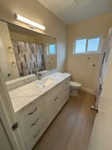 A modern bathroom renovation by TG Renovations showcasing a white vanity with a marble-look quartz countertop, a large mirror with a contemporary vanity light, and light wood-look flooring.