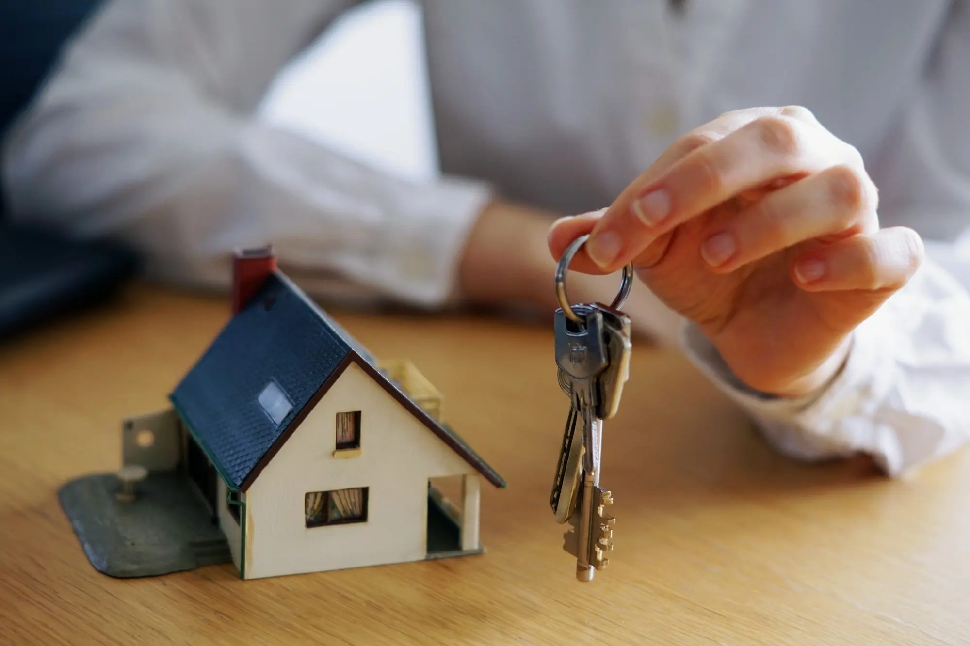 Hand holding house keys next to a miniature house model, symbolizing renting an ADU to family members – TG Renovations