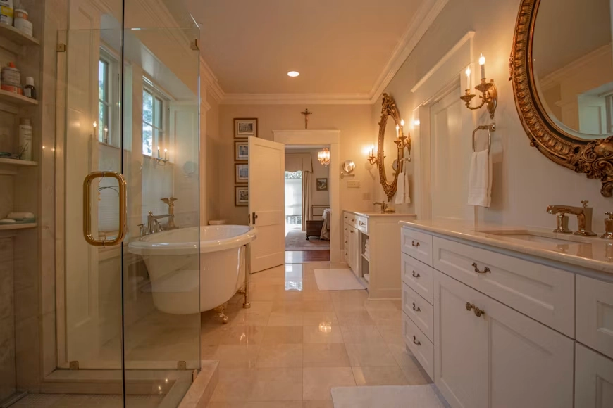 A photograph of an elegant, traditional bathroom remodel by TG Renovations. The spacious layout features a glass-enclosed shower with brass hardware, a classic freestanding clawfoot bathtub, and polished tile flooring. To the right, dual white vanities with marble countertops are accented by ornate gold mirrors and classic wall sconces, with an open door in the background leading to an adjoining bedroom.