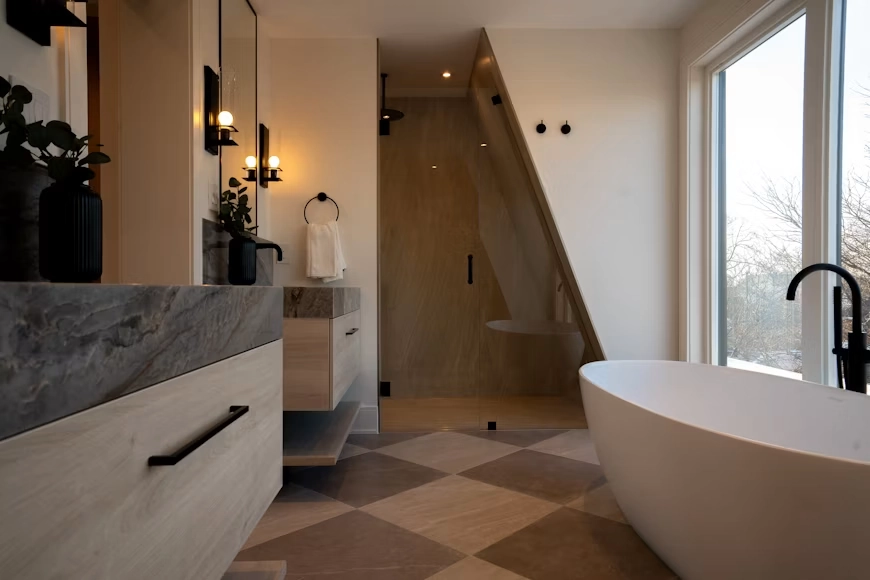 A complex, modern bathroom renovation by TG Renovations featuring a custom glass-enclosed shower built into a slanted structural ceiling. The space includes a freestanding white tub near a large window, a floating wood vanity with a thick marble countertop, matte black fixtures, and geometric floor tiles. This architectural transformation illustrates the type of extensive plumbing and framing work discussed in 'Legal and Structural Realities: Do I Need a Permit to Remodel My Bathroom?'.