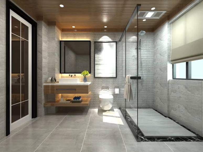 A photograph of a high-end, modern bathroom renovation by TG Renovations, showcasing a sleek floating vanity with a vessel sink and a large, backlit mirror, adjacent to a wall-hung toilet. The space features a glass-enclosed walk-in shower area with a rainfall head and a slatted wood floor mat, set against grey marble-tiled walls and floor. The design is complemented by a warm wood plank ceiling with recessed lighting and abstract wall art.