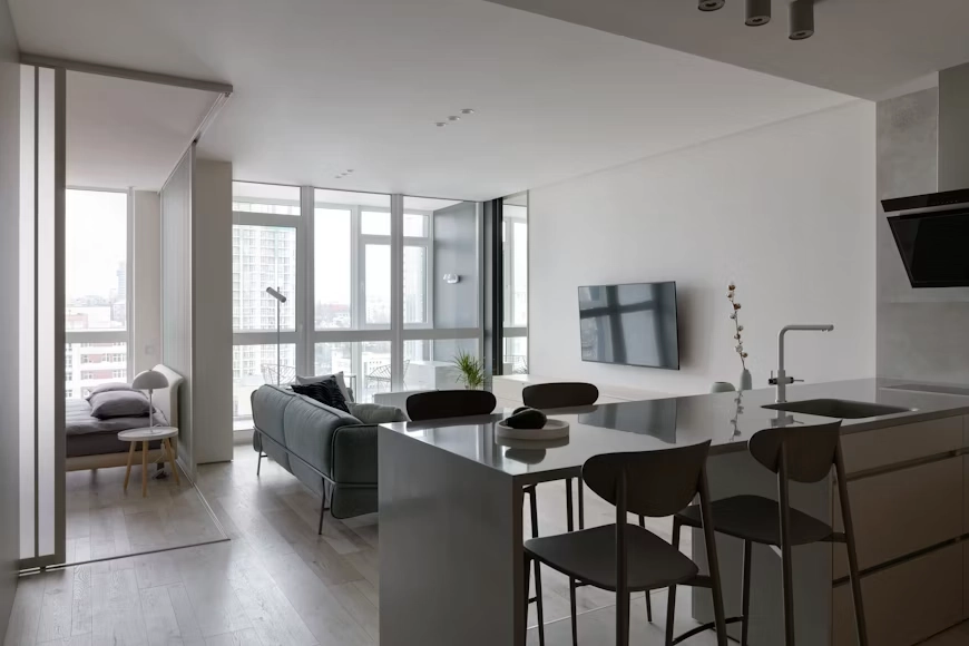 A modern, minimalist open-concept apartment renovation by TG Renovations, featuring a sleek grey kitchen island, a comfortable living area with city skyline views, and a sliding glass-partitioned bedroom. This sophisticated space perfectly illustrates the transformative design possibilities discussed in 'The Elite Urban Blueprint: Can You Renovate An Apartment?'.