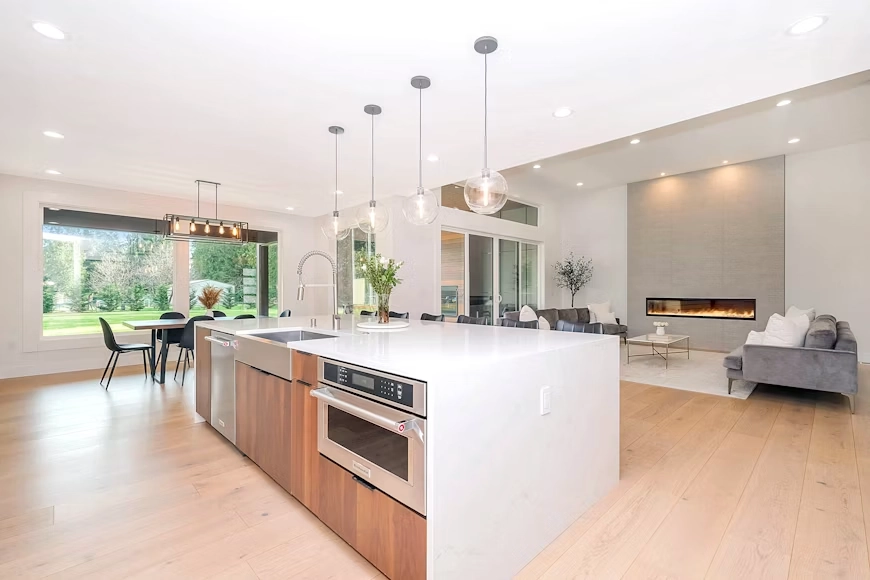 A bright, open-concept home remodel by TG Renovations, featuring a modern kitchen with a large white island, wood cabinetry, stainless steel appliances, and glass pendant lights. The kitchen flows seamlessly into a dining area with large windows and a living room centered around a sleek linear fireplace and grey sofas, all unified by light wood flooring.