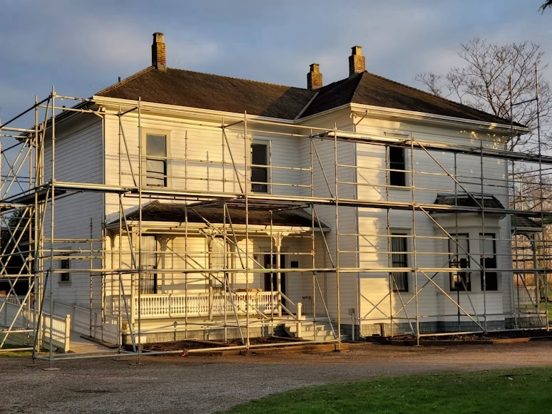 A large two-story white house surrounded by extensive metal scaffolding during an exterior renovation by TG Renovations. The property is bathed in warm sunlight, illustrating the scale of whole-home remodeling projects for 'The Premium Guide: How Much Does a Home Renovation Cost?'.